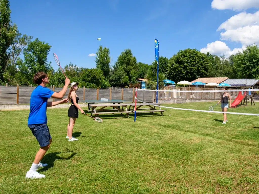 Amis jouant au badminton sur l'aire de jeux du camping Saint-Martin à Sorèze, proche d'Albi, sous un ciel ensoleillé et entouré de nature.