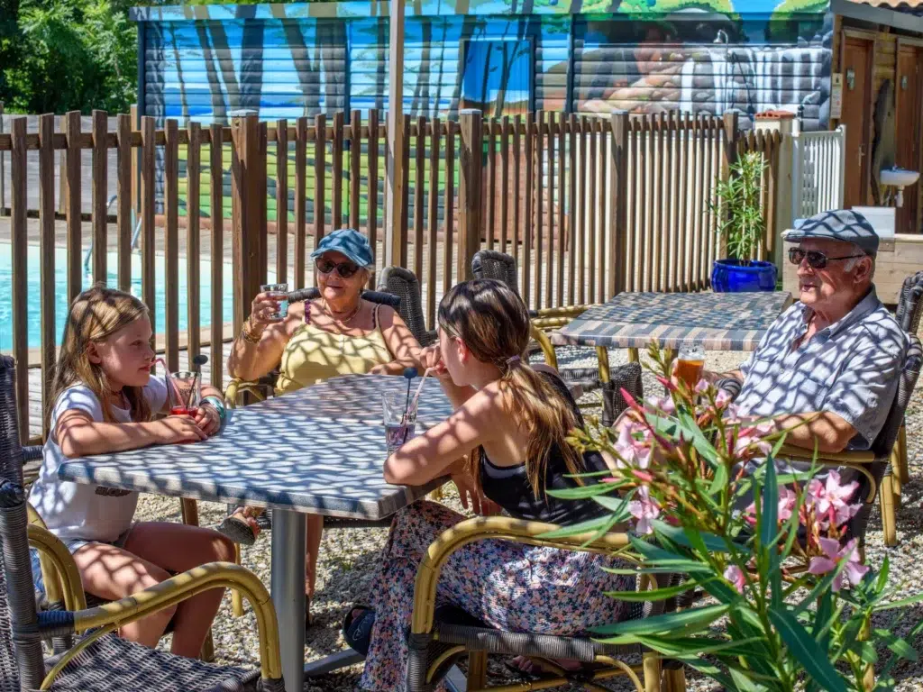 Famille en détente sur la terrasse ombragée du snack-bar du camping Saint-Martin, dans le Tarn, avec vue sur la piscine et ambiance conviviale.