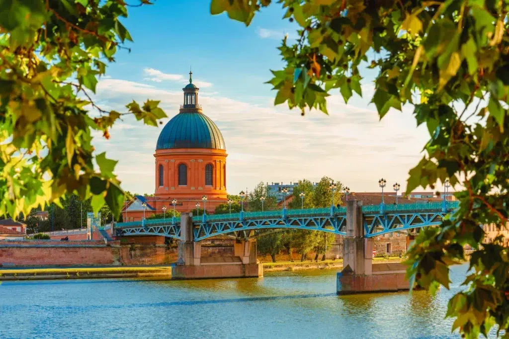 Le dôme de la Gravette et le pont sur la Garonne à Toulouse, encadré par des feuilles vertes, mettant en valeur le patrimoine architectural de la ville.