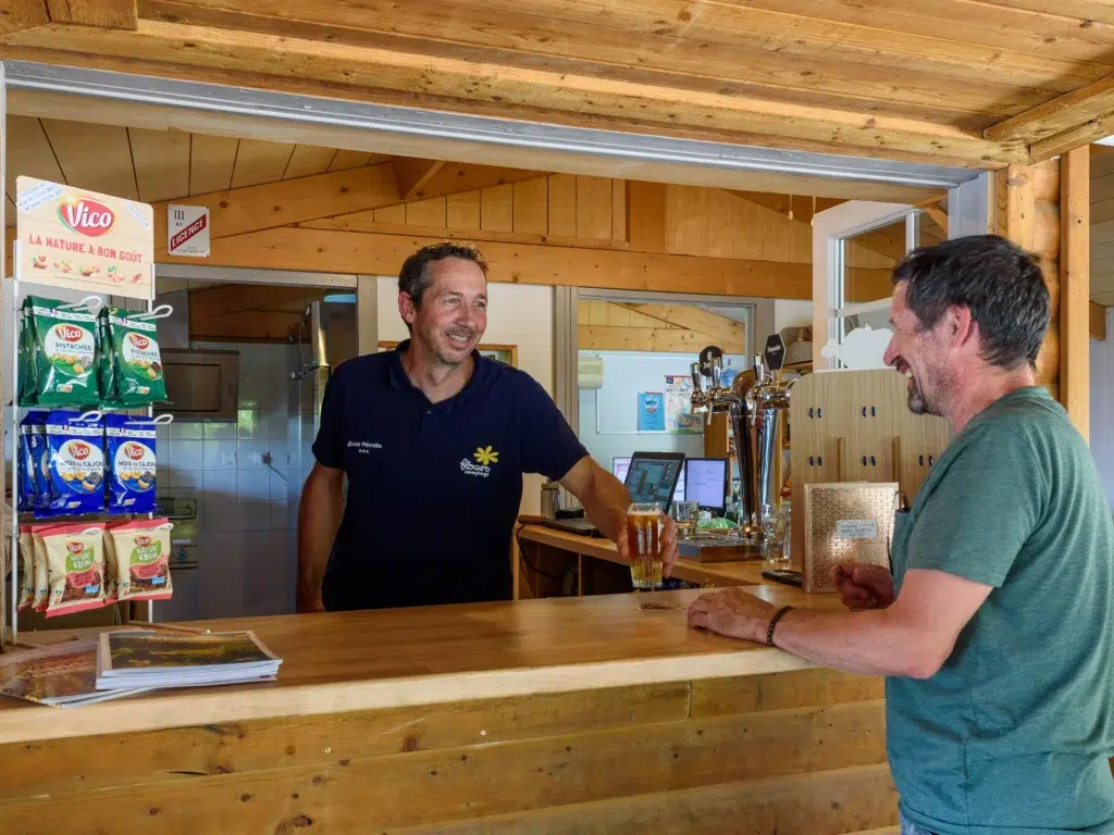 Snack/bar convivial au Flower Camping Saint Martin avec un employé souriant servant un client au comptoir en bois. Ambiance chaleureuse et produits locaux pour des moments de détente en camping.