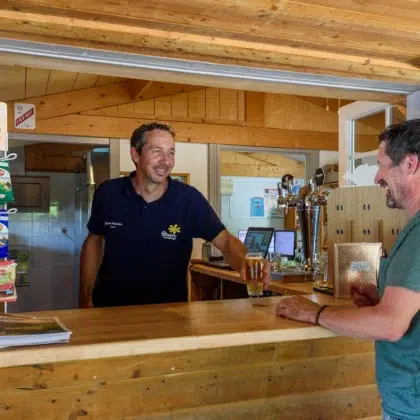 Accueil 20 Snack/bar convivial au Flower Camping Saint Martin avec un employé souriant servant un client au comptoir en bois. Ambiance chaleureuse et produits locaux pour des moments de détente en camping. Détendez-vous au snack/bar de notre Flower Camping Saint Martin, où l'accueil chaleureux et les produits locaux vous garantissent des moments inoubliables.